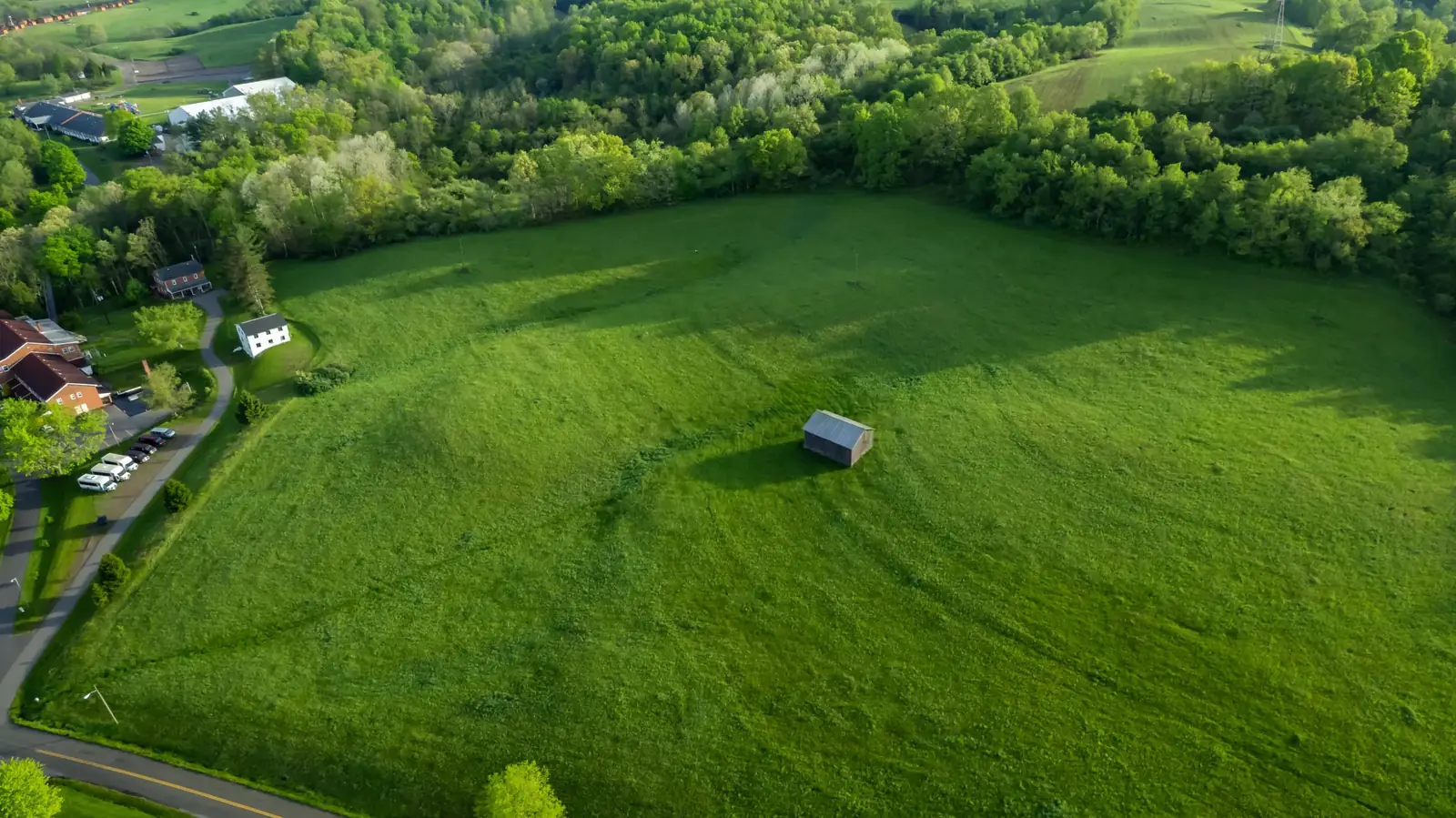 Aerial photo of a hay field: a tree line along the top, a shed in the middle of the field, two power poles in the grass, a white farmhouse and a road down the left side.